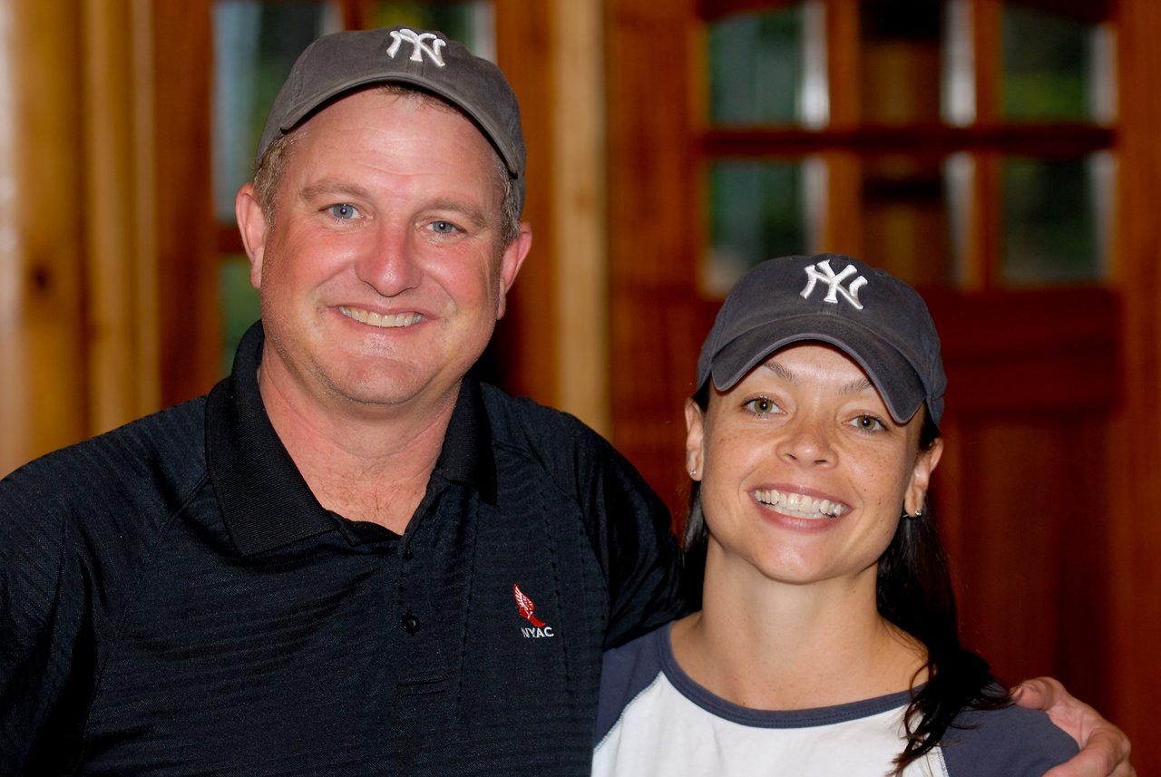 Two people wearing New York Yankees caps smile while posing together indoors.
