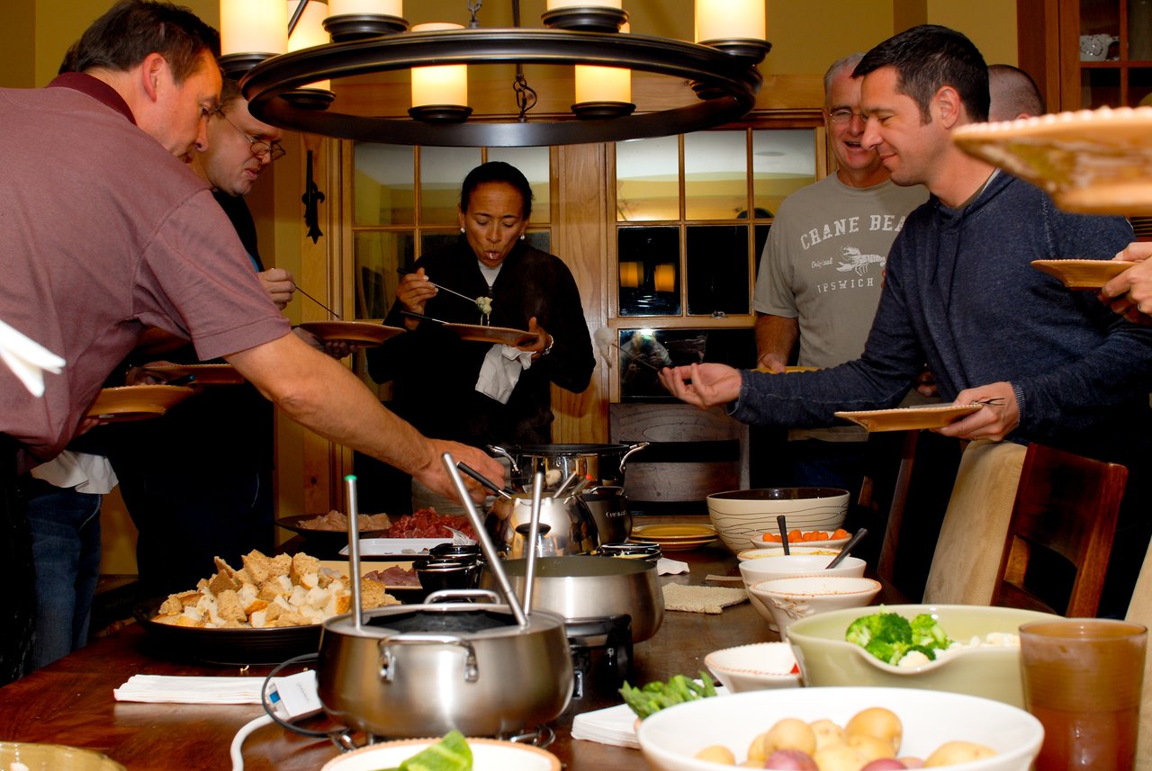 A group of people gathers around a table, serving themselves food from a fondue spread.