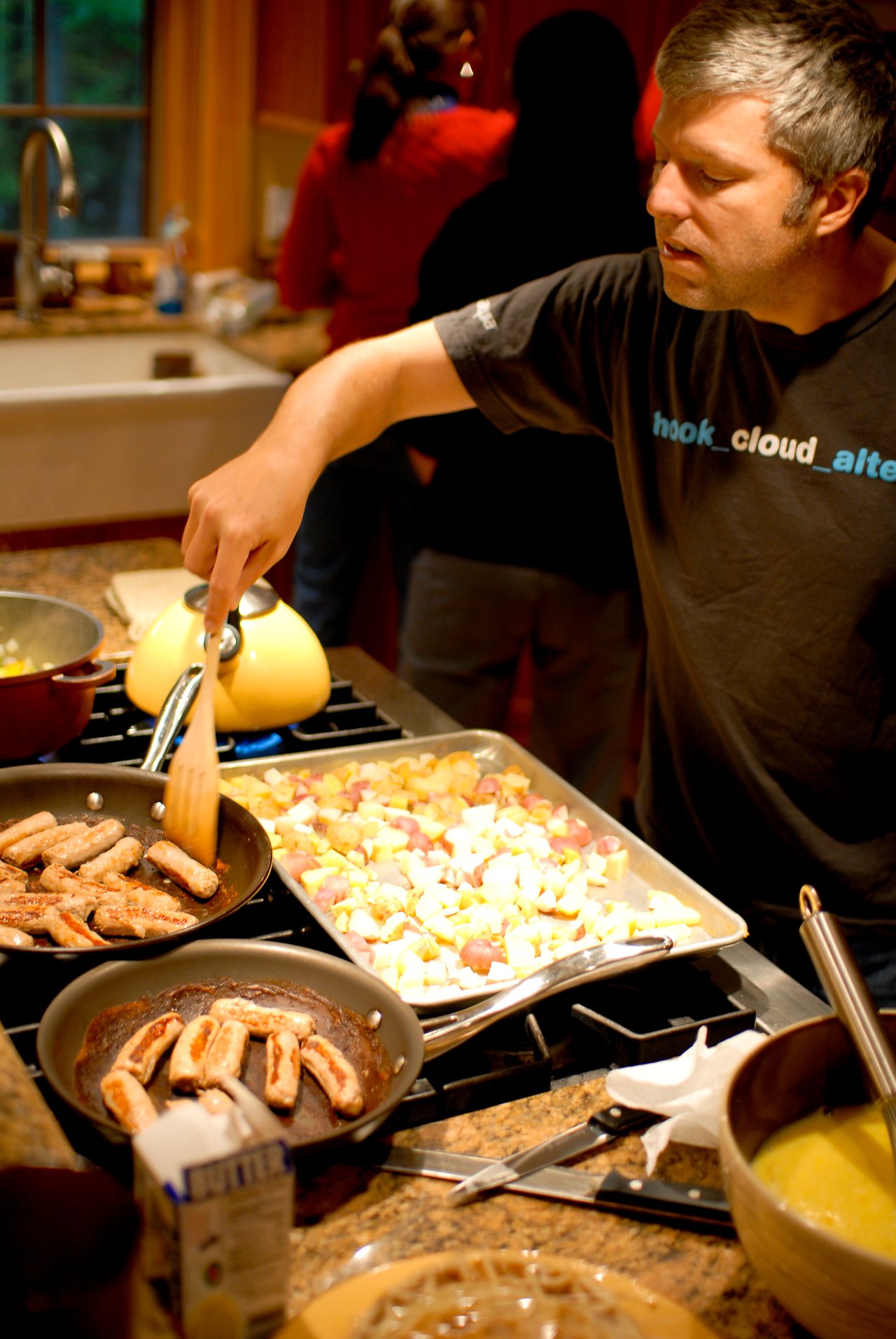 A man cooks sausages in a pan while roasting potatoes on a baking sheet in a kitchen.