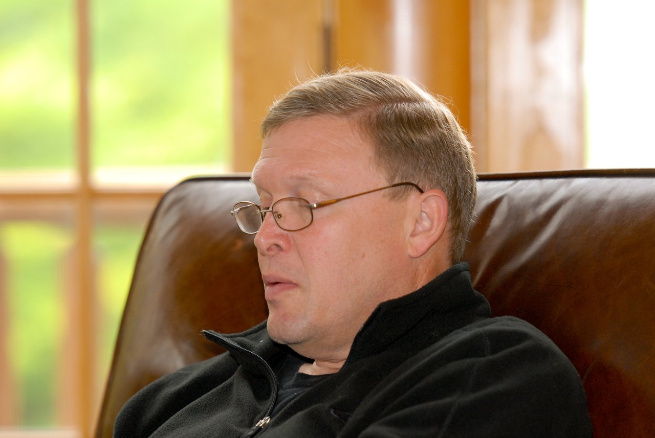 A man wearing glasses and a black jacket sits on a leather chair, looking down with a focused expression.