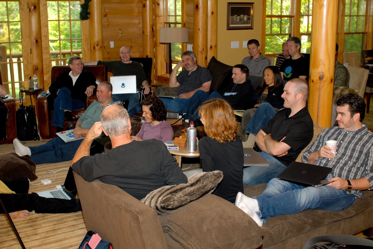 A group of people in a cozy cabin setting, sitting with laptops and drinks, engaged in a discussion.