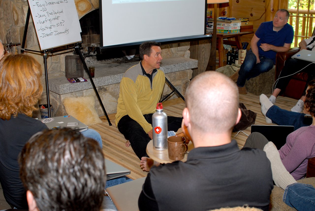 A man sits on the floor leading a discussion while a group of people listens in a casual setting.