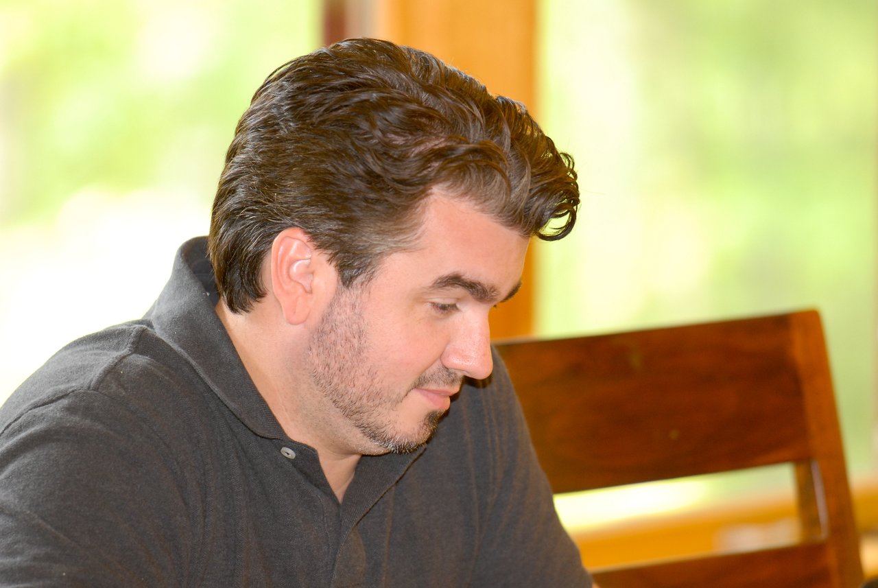 A man in a dark polo shirt looks down with focus, sitting indoors near a wooden chair.