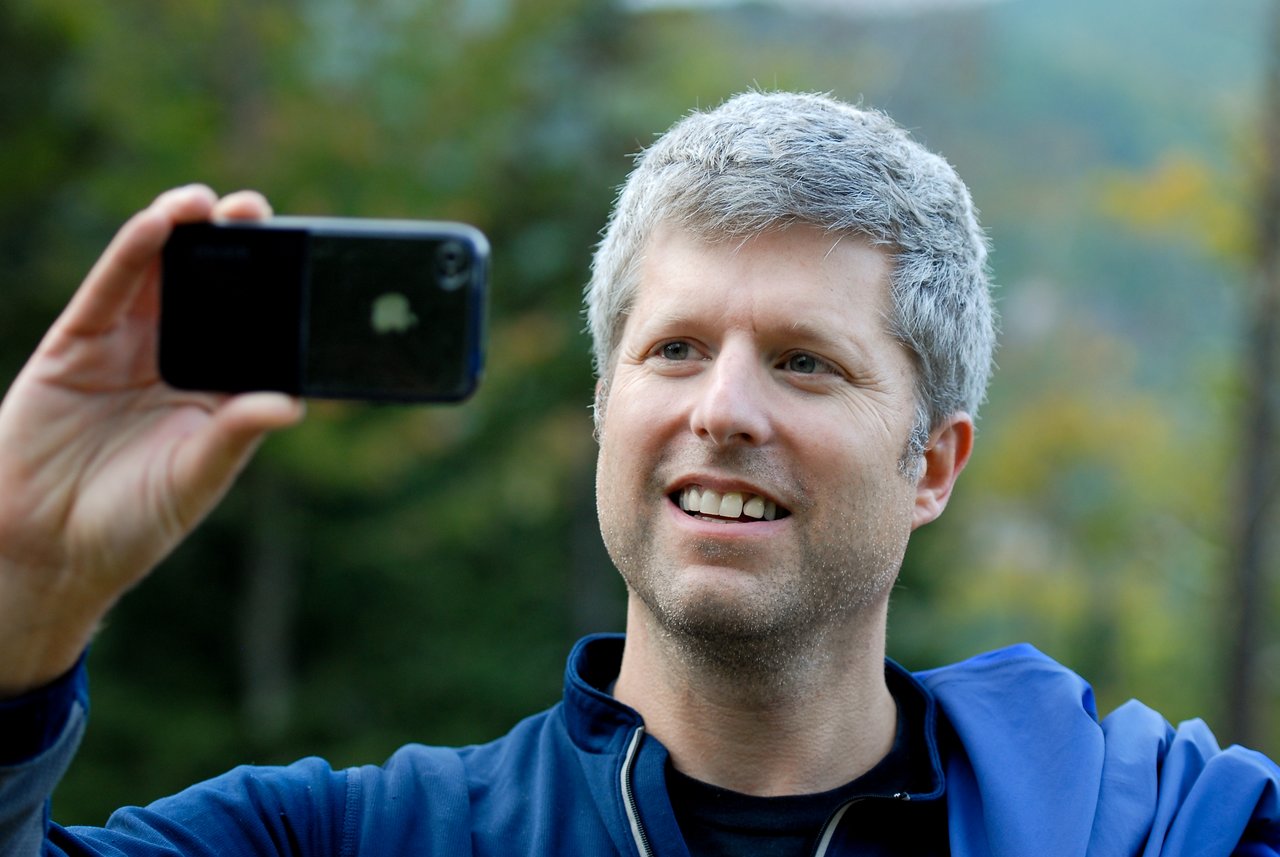A man in a blue jacket smiles while holding up a smartphone to take a photo or video.