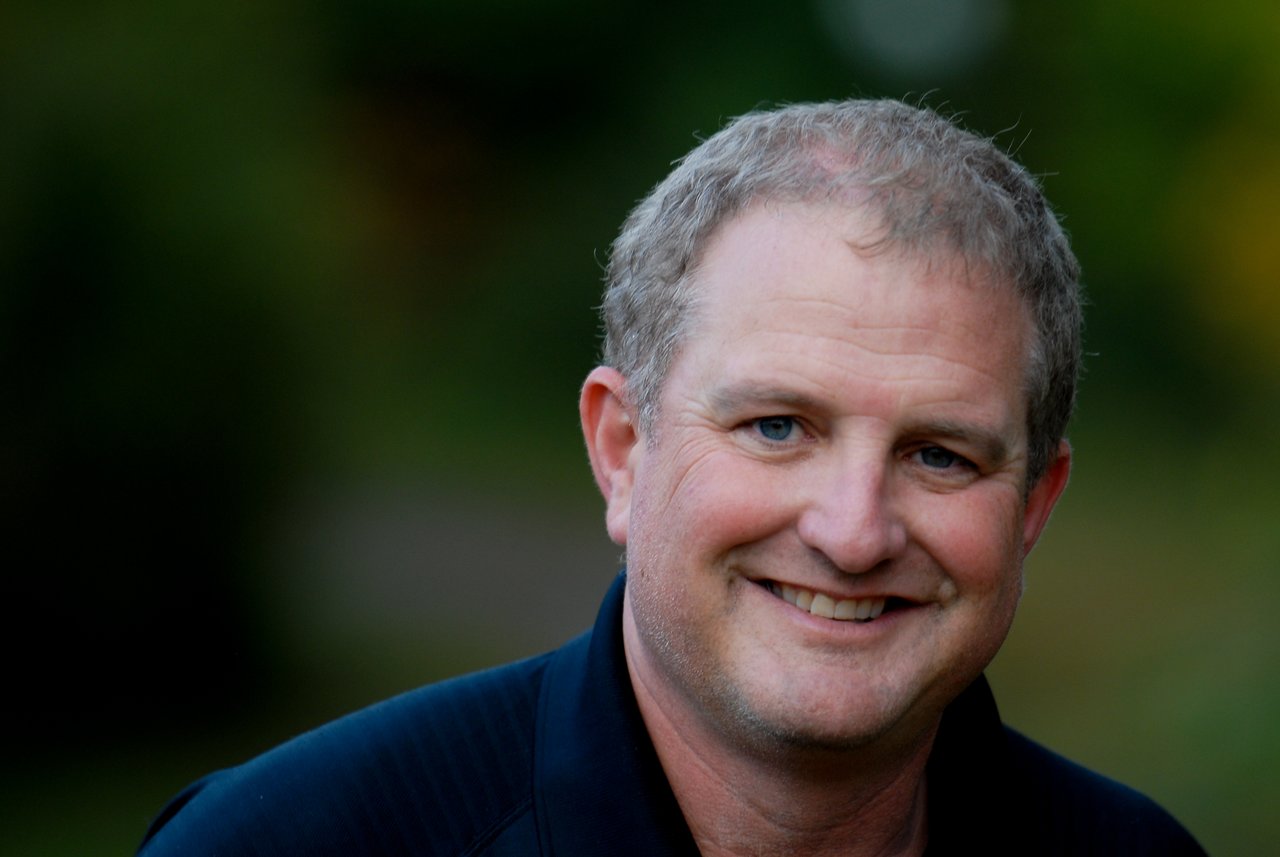 A man wearing a dark shirt smiles at the camera with a blurred green background.