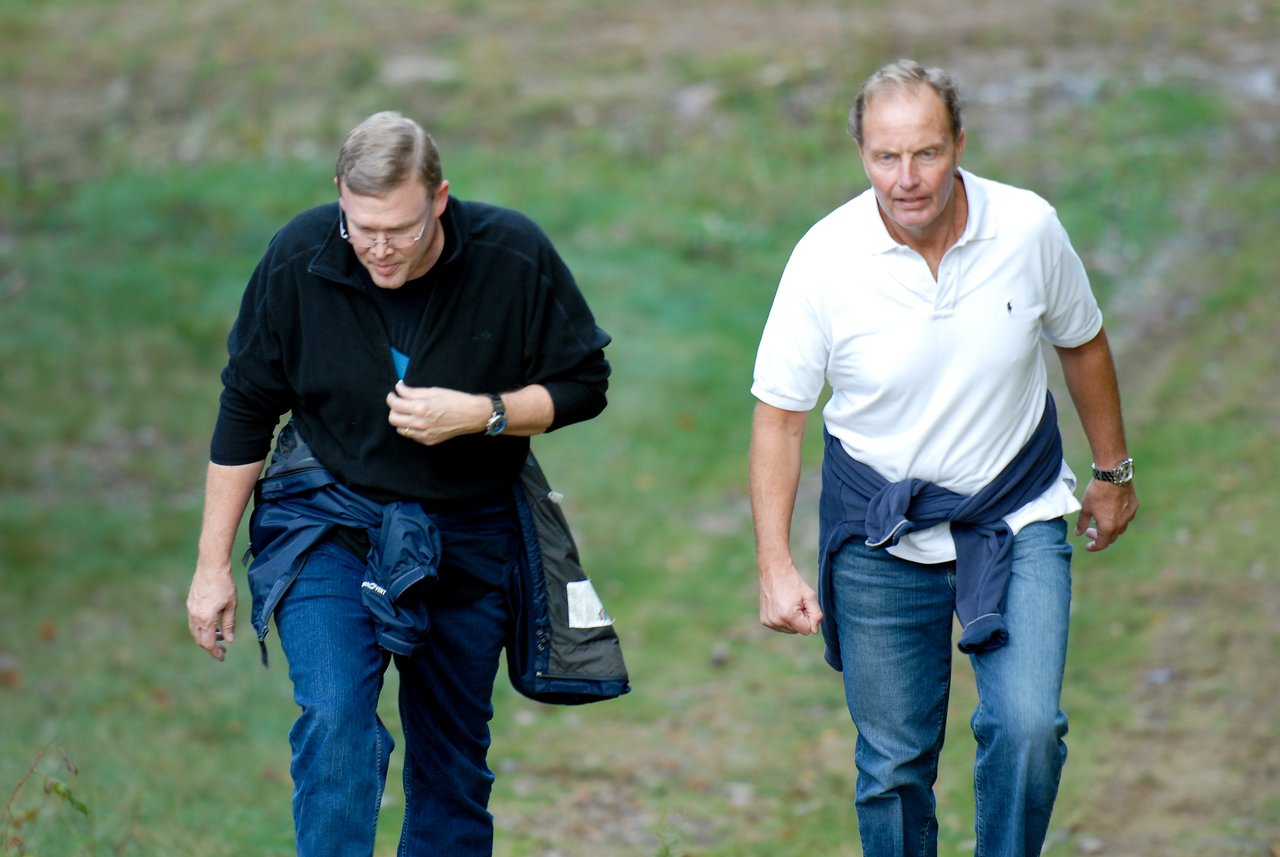 Two men walk uphill on a grassy path, both wearing jackets tied around their waists.