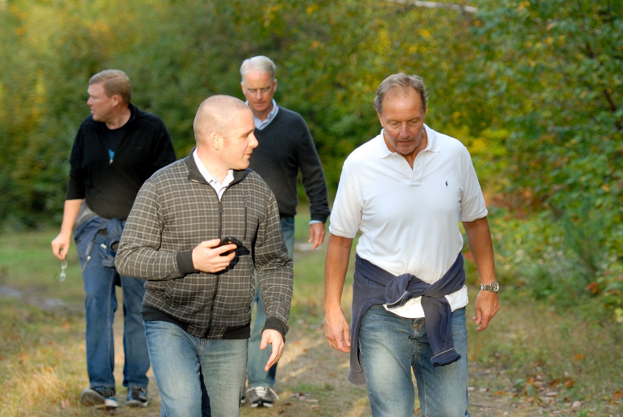 Four men walk along a grassy trail, with two in front engaged in conversation.