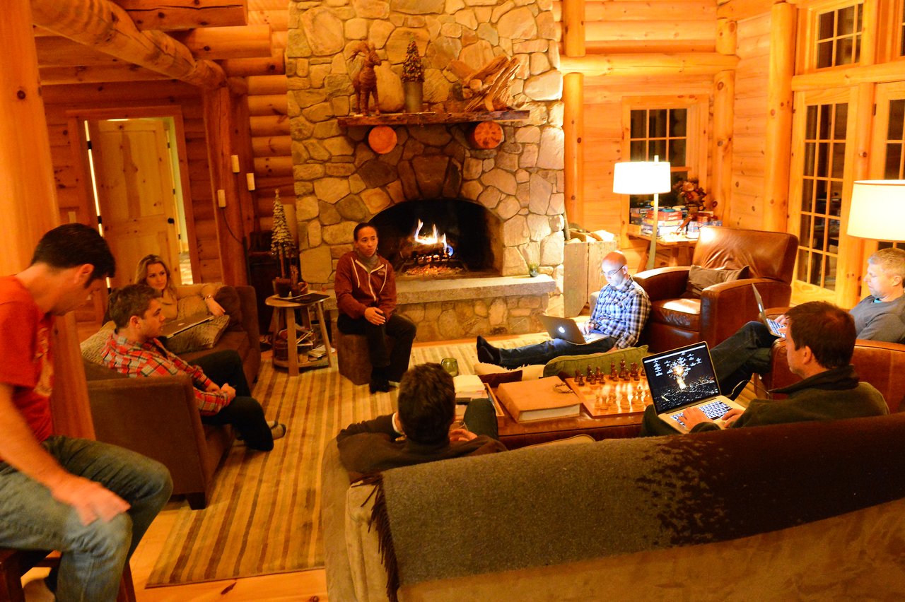 A group of people in a log cabin, some working on laptops, while one person speaks near a fireplace.