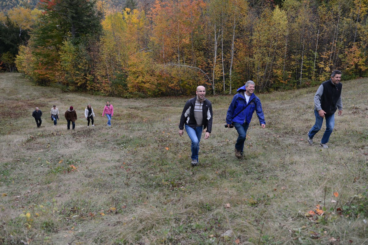 A group of people hikes up a grassy hill surrounded by autumn trees during an outdoor company event.