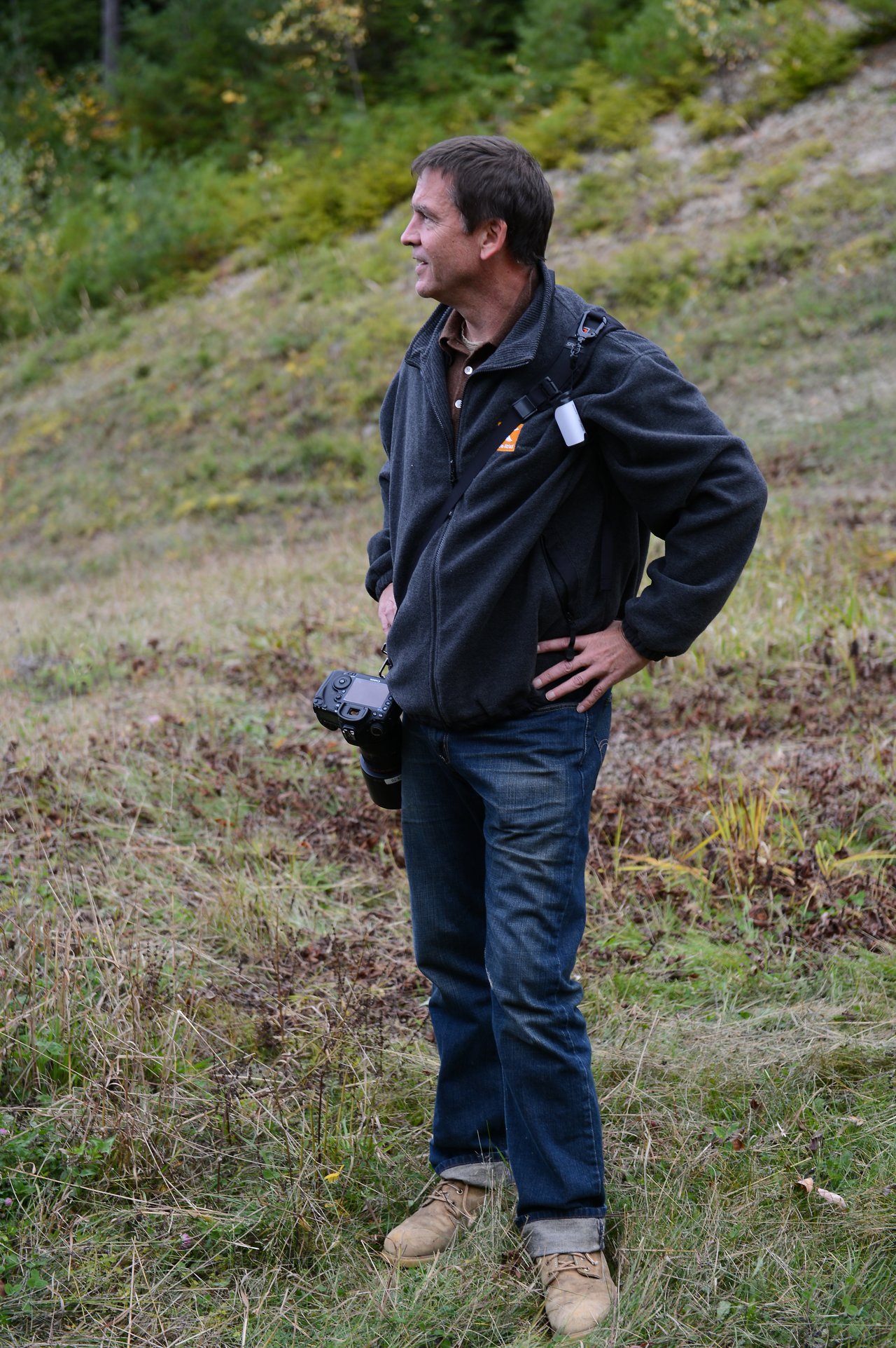 A man in a jacket stands on a grassy hillside, holding a camera and looking to the side.