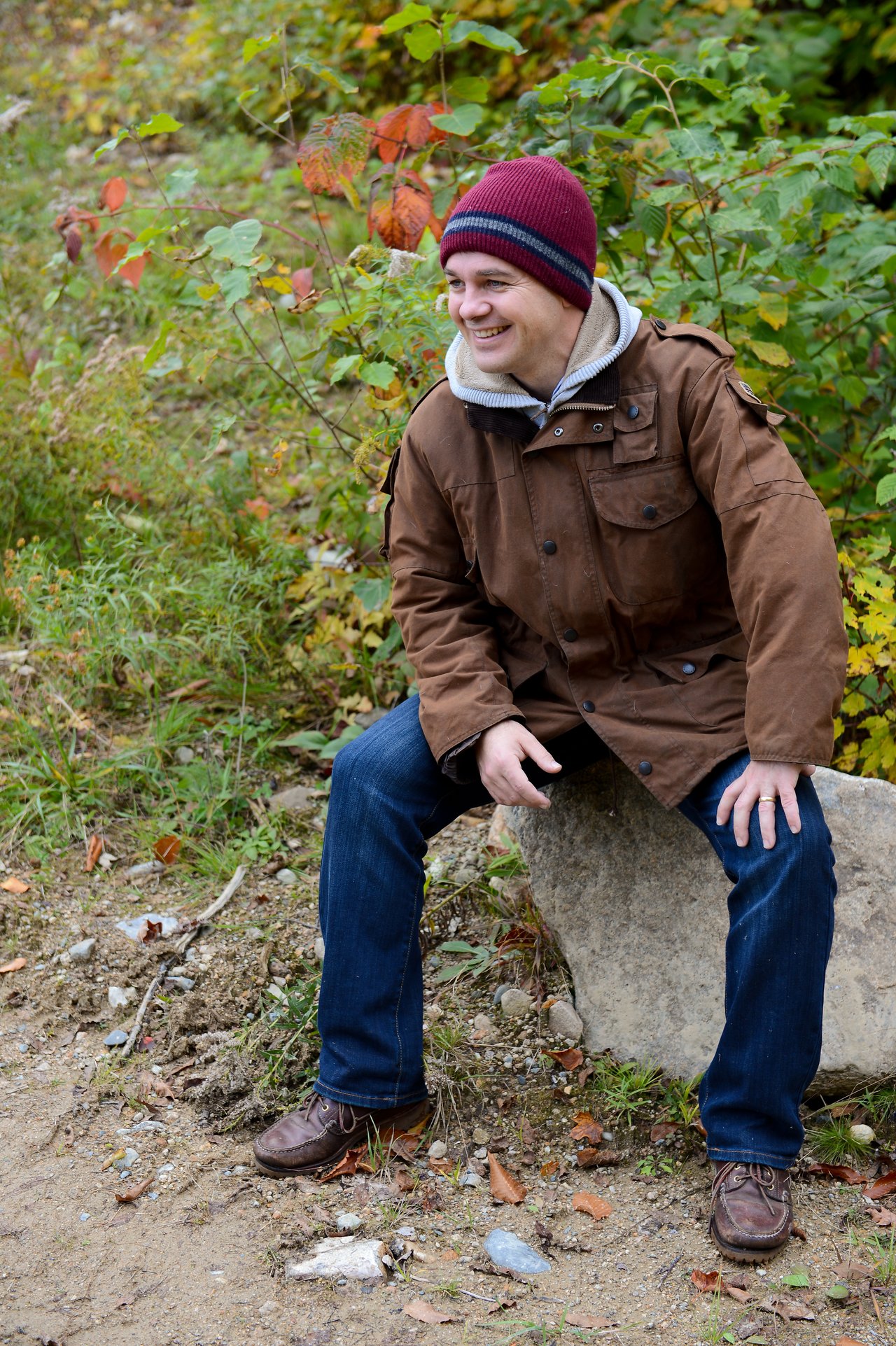 A man in a brown jacket and beanie sits on a rock outdoors, smiling and looking to the side.