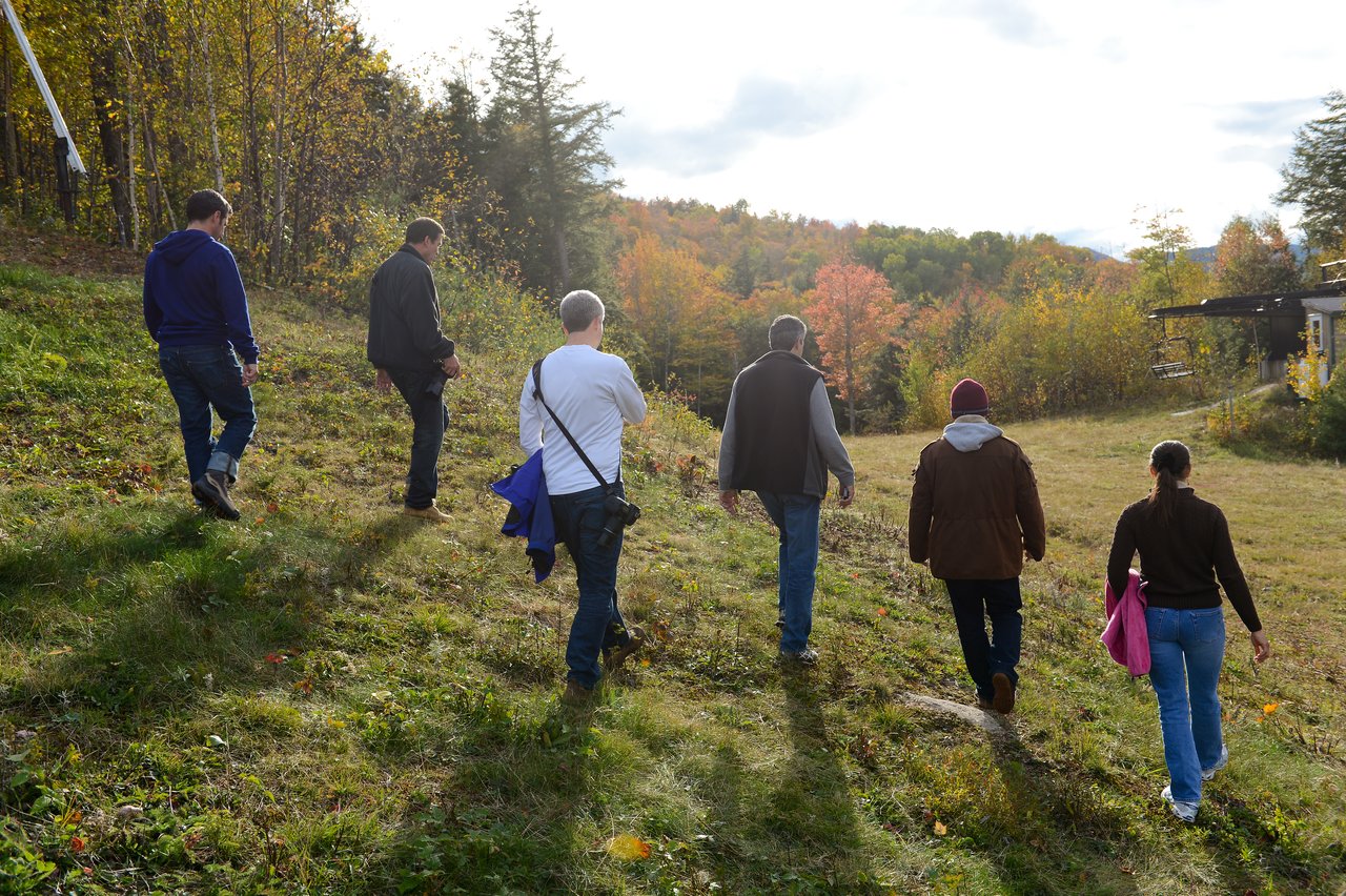 A group of six people walks uphill on a grassy path surrounded by autumn trees.