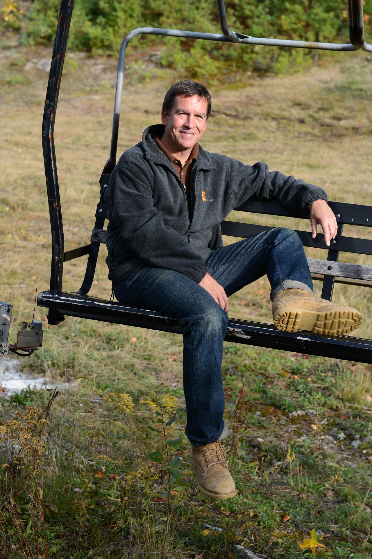 A man in a jacket and boots sits on a ski lift, smiling at the camera.