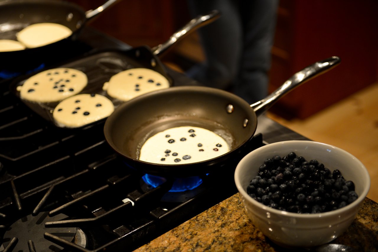 Blueberry pancakes cooking on a stovetop, with a bowl of fresh blueberries nearby.