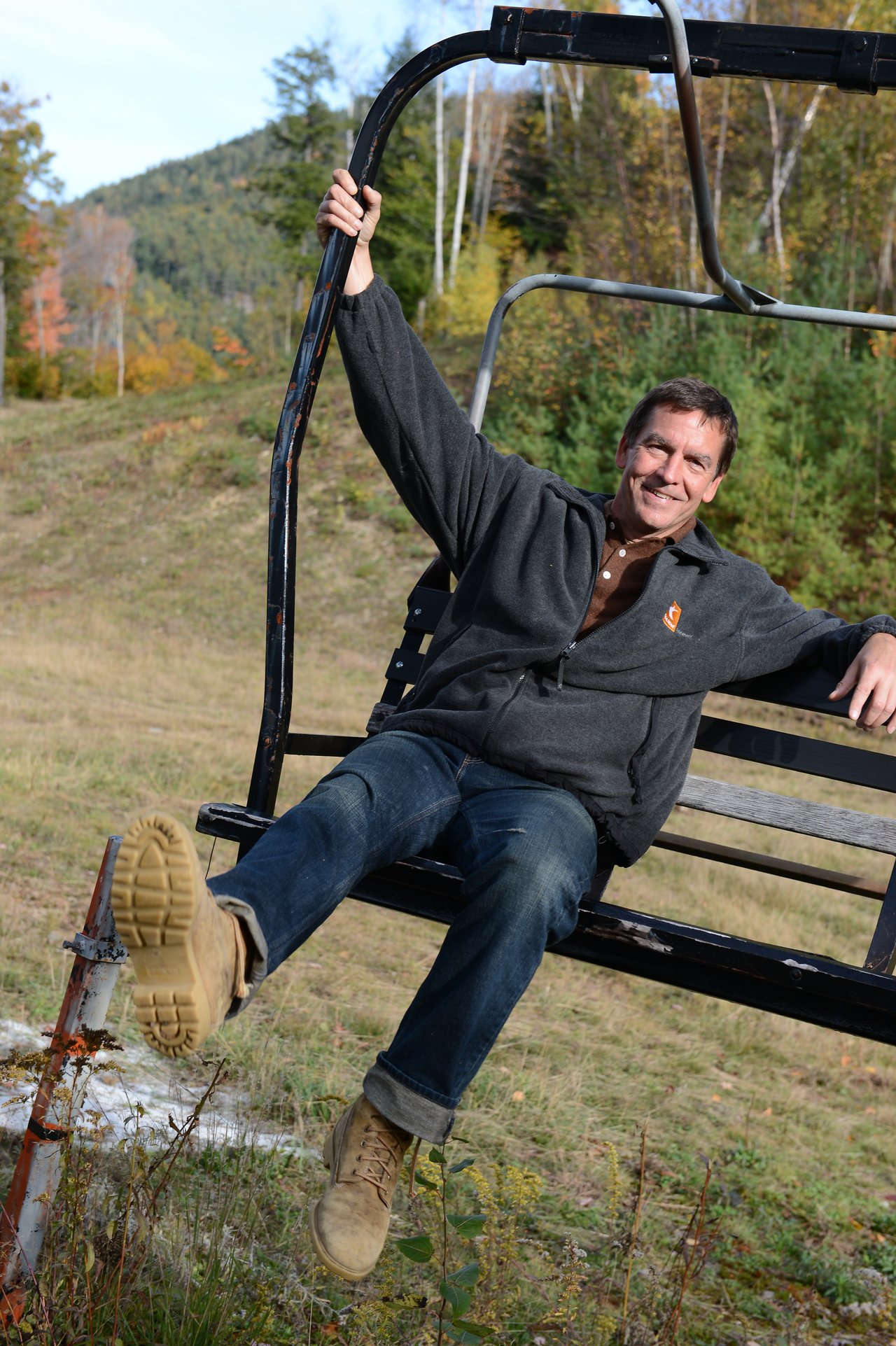 A man in a dark jacket sits on a ski lift, smiling and playfully holding onto the metal frame.