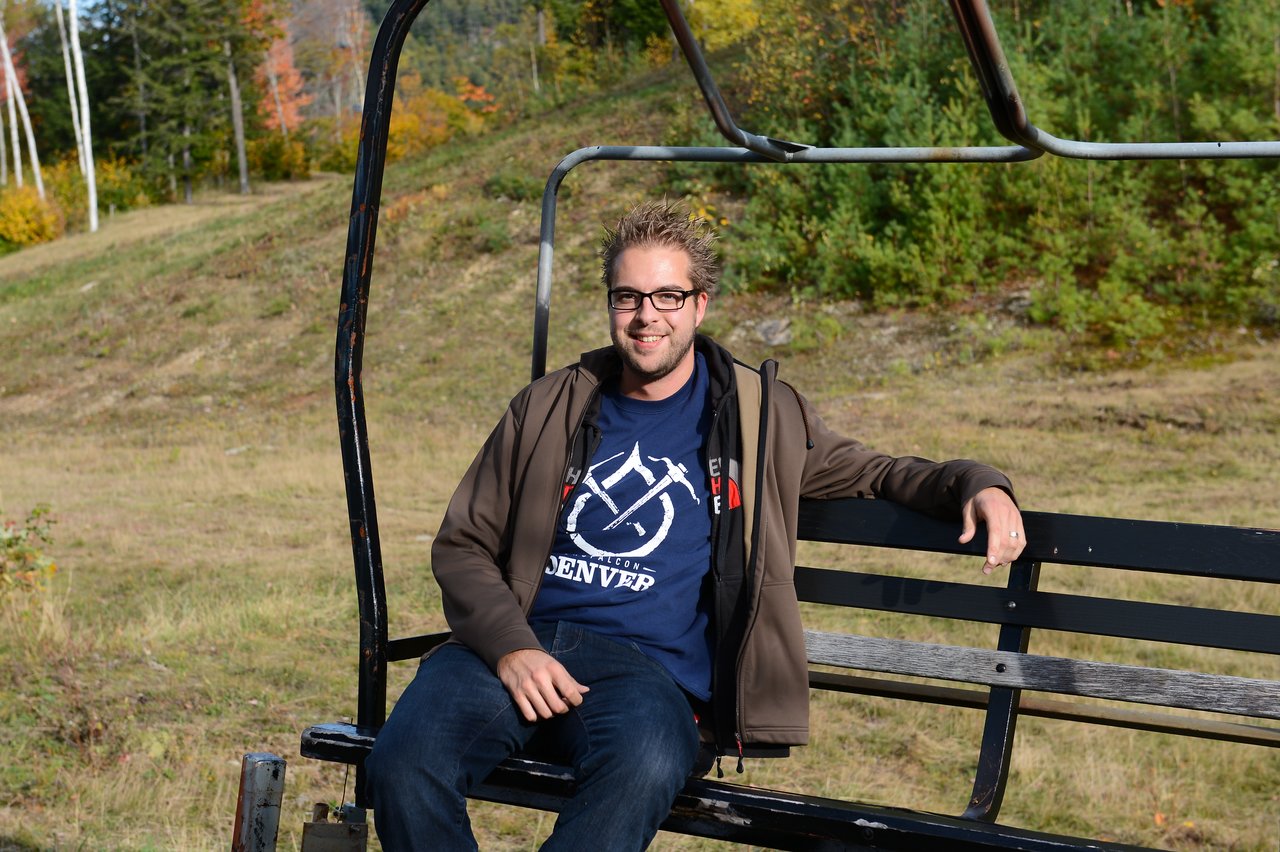 A man wearing glasses and a jacket sits on a ski lift bench, smiling at the camera.