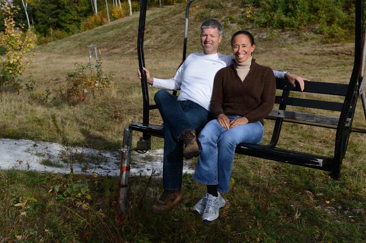 Two people sit on a stationary ski lift chair outdoors, smiling at the camera.