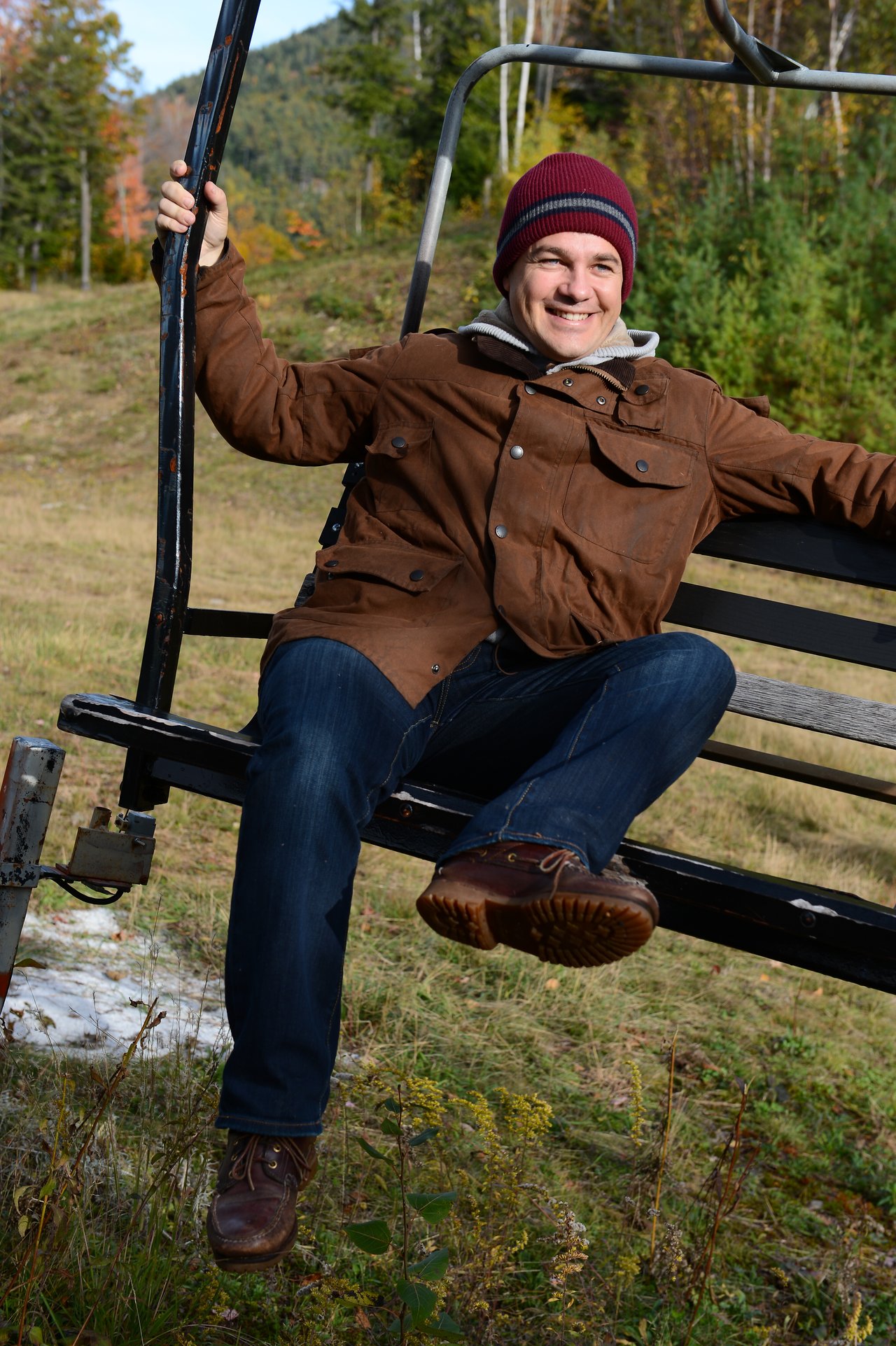 A smiling person in a brown jacket and red beanie sits on a ski lift, holding onto the frame.