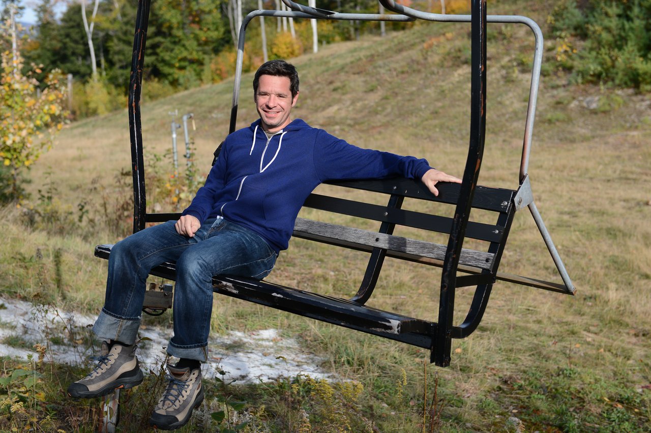 A man in a blue hoodie sits and smiles on a stationary ski lift chair in an outdoor setting.