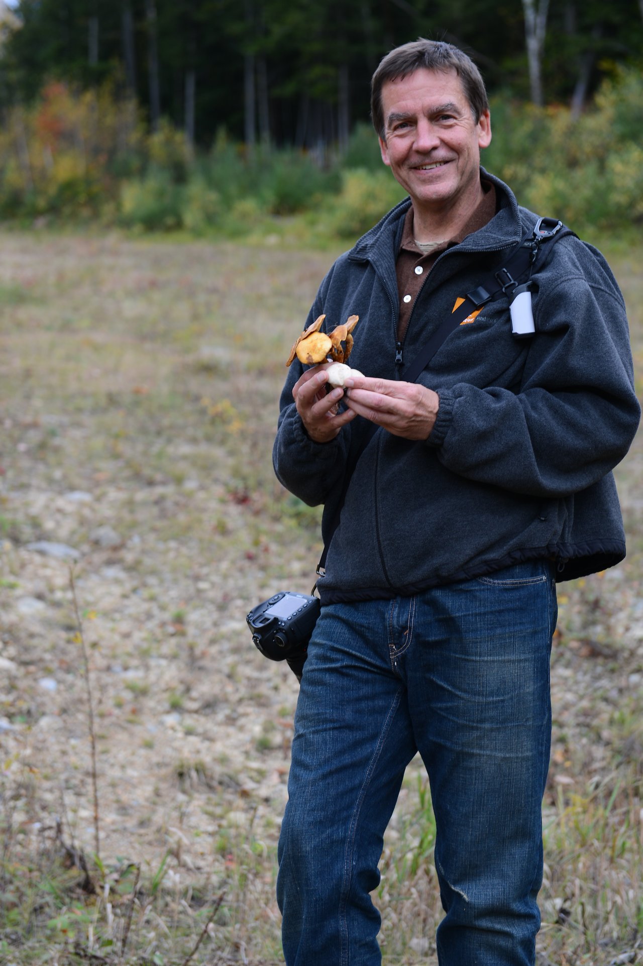 A man standing outdoors holds mushrooms in his hands and smiles, with a camera hanging from his waist.