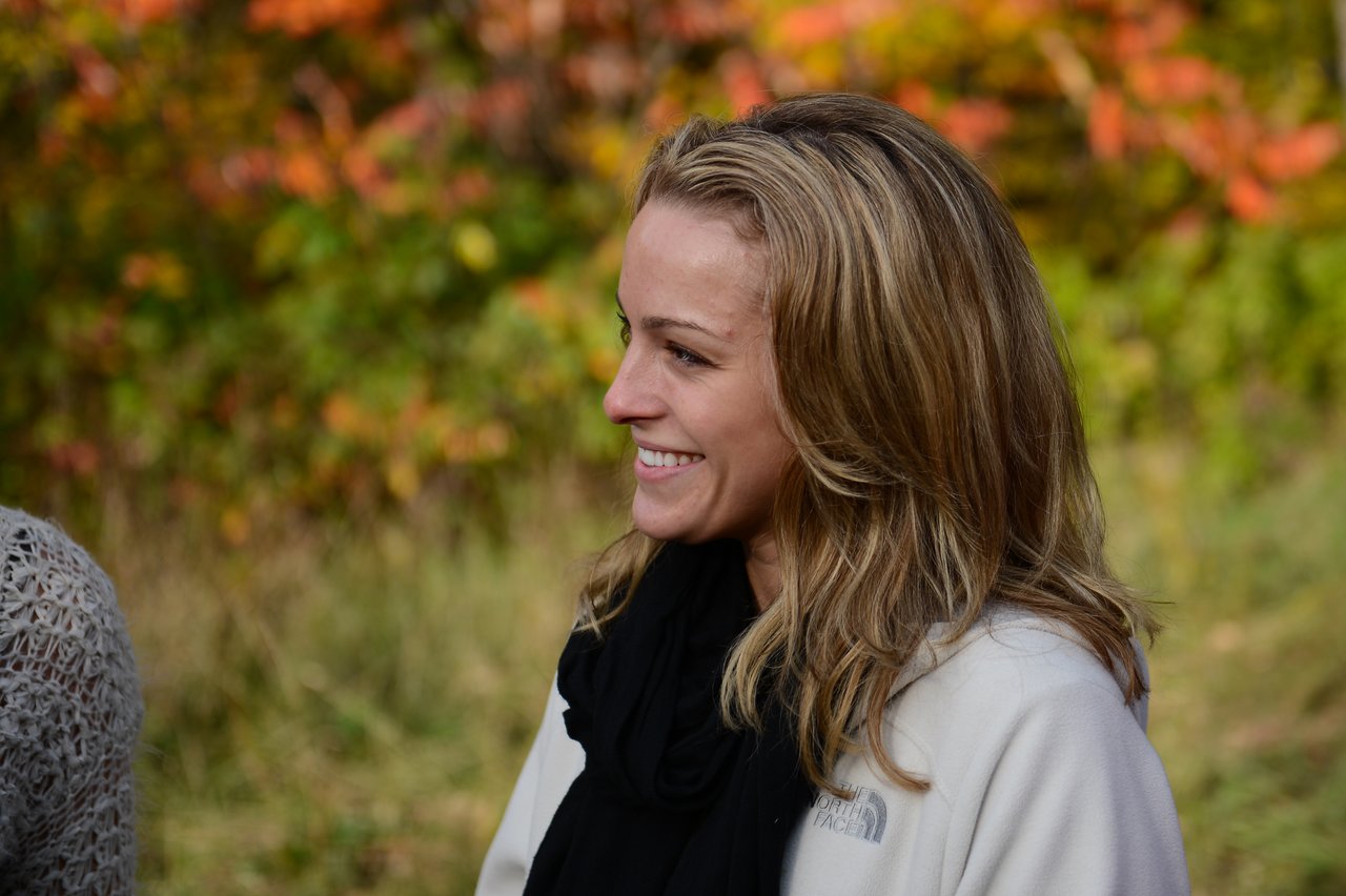 A woman in a white jacket smiles while talking to someone outdoors with autumn foliage in the background.