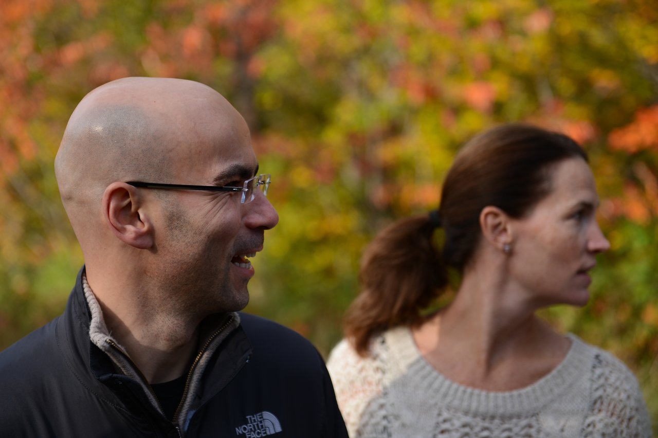 A man in a black jacket smiles while a woman in a white sweater looks away in an outdoor setting.