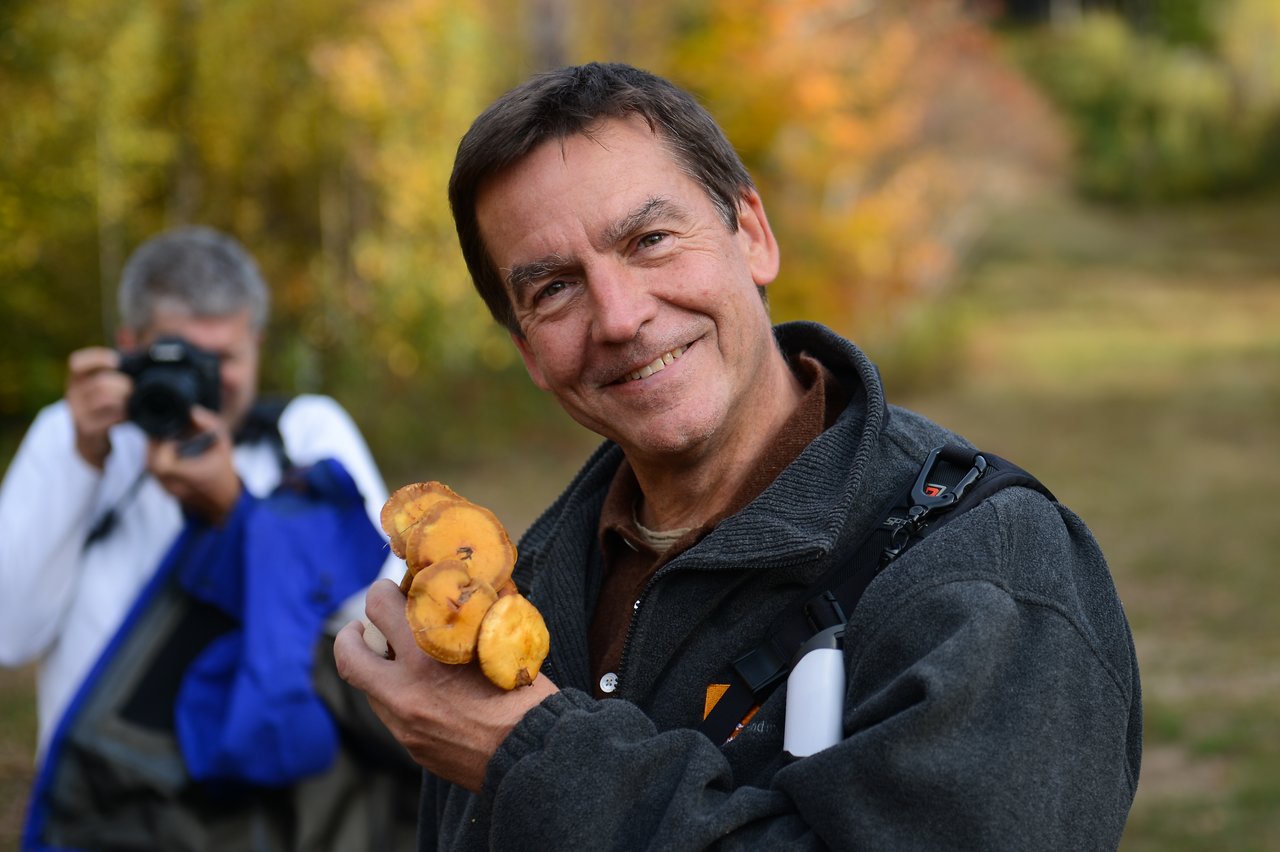 A man smiles while holding a cluster of mushrooms, with another person in the background taking a photo.