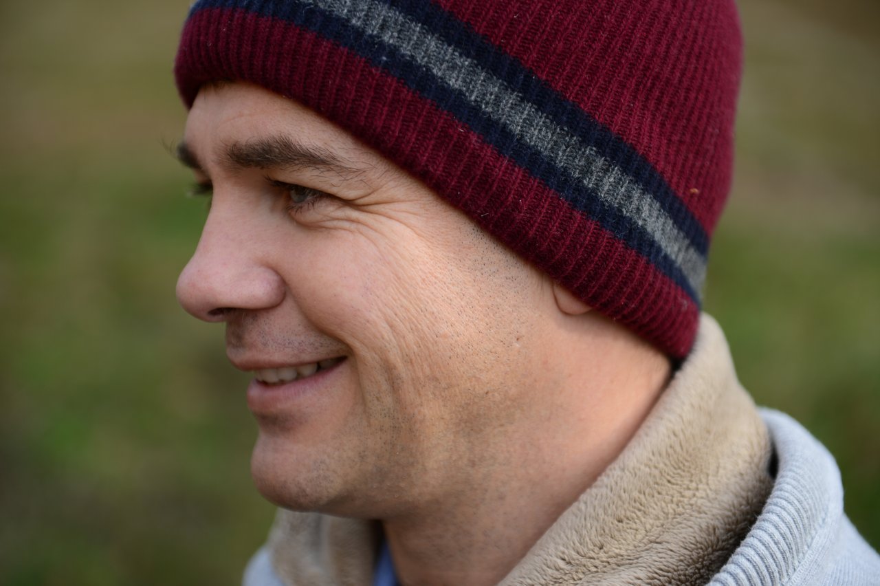 Close-up of a person wearing a red and gray beanie, smiling while looking to the side outdoors.