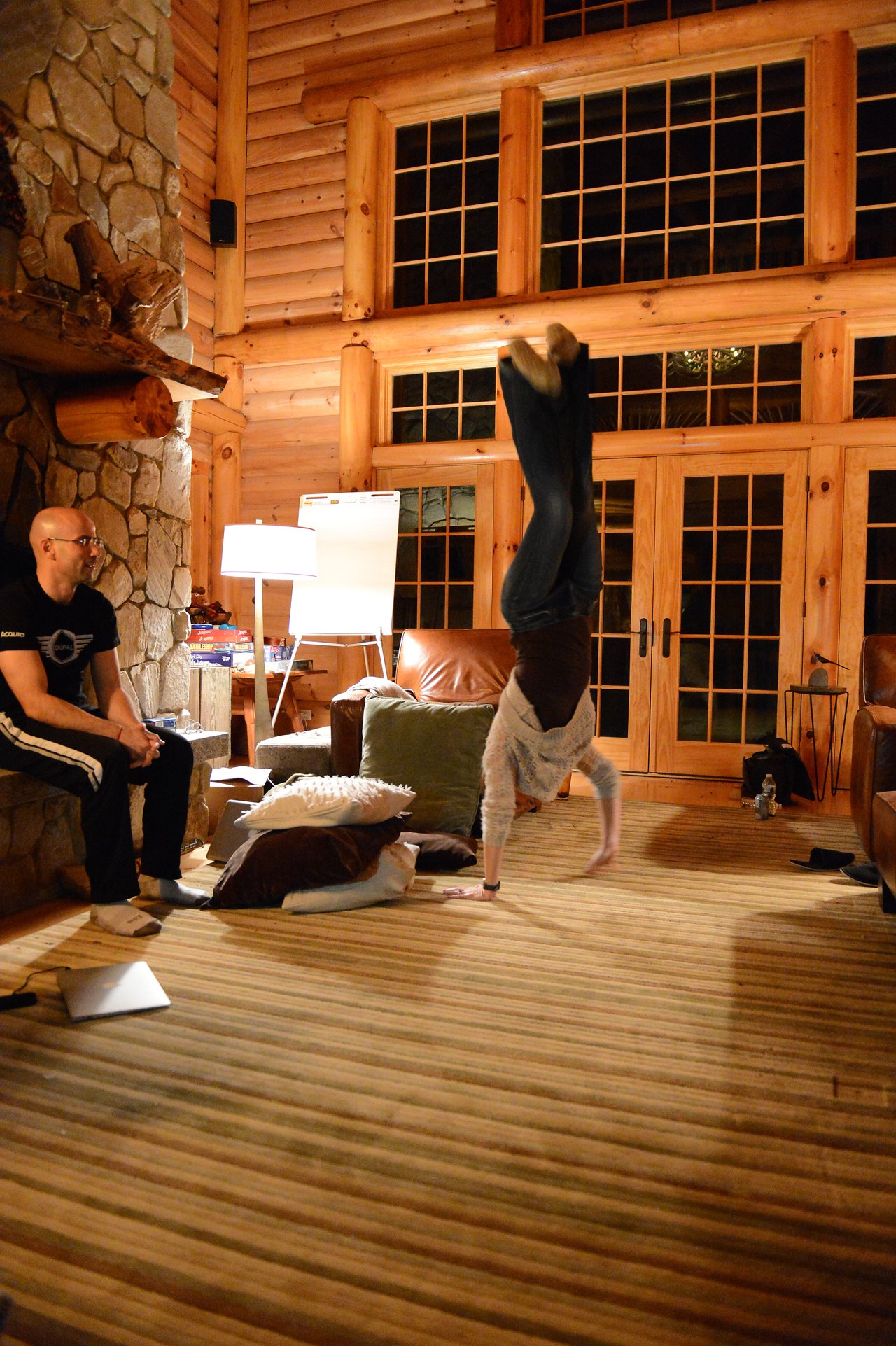 A person does a handstand in a cozy wooden cabin while another person watches and smiles.