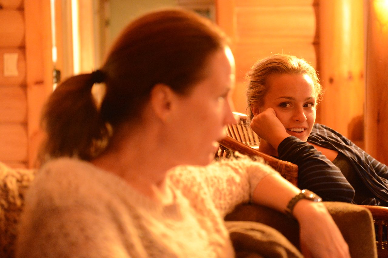 One woman speaks while another listens and smiles in a warmly lit indoor setting.
