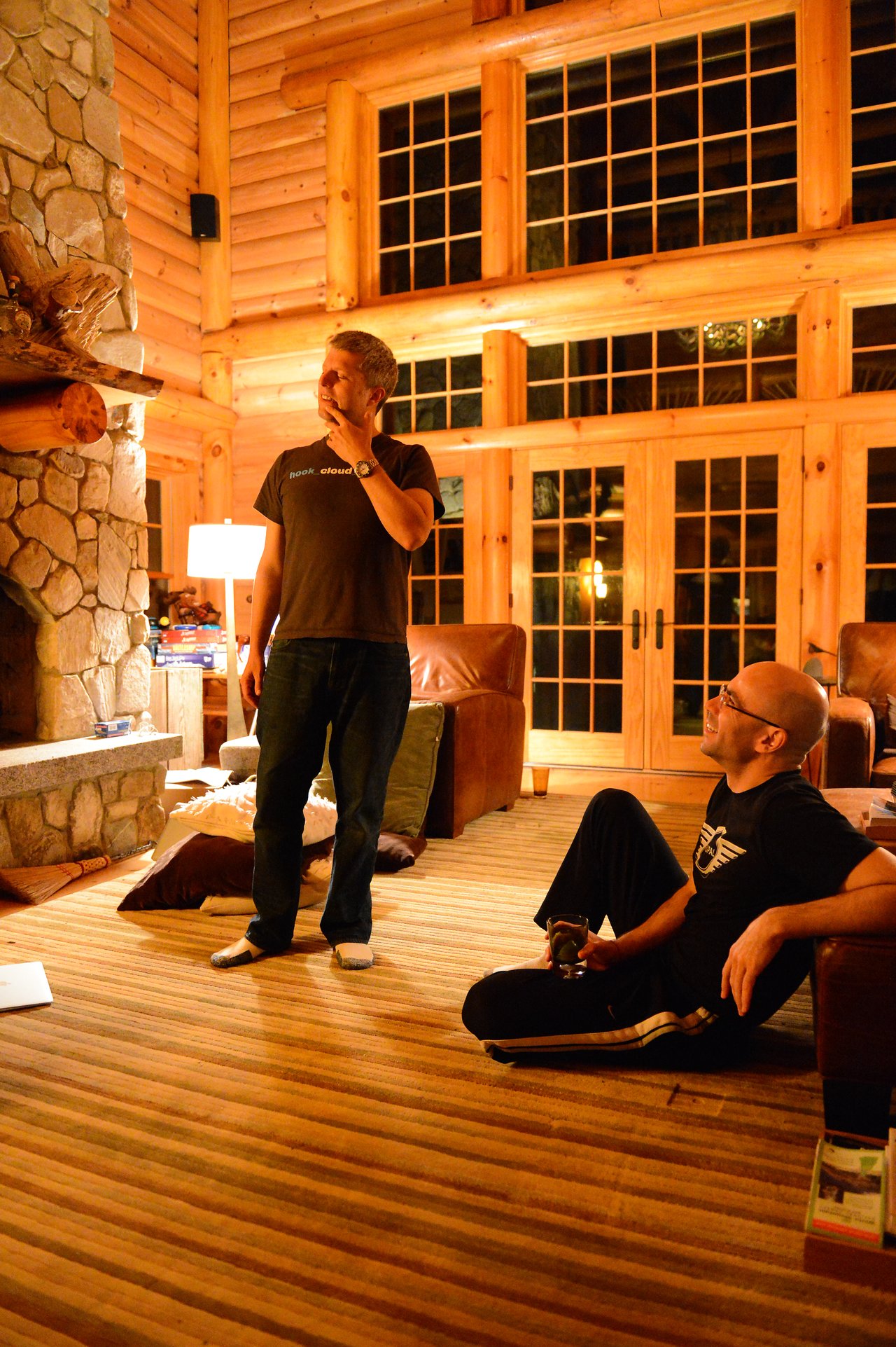 A man stands and gestures while another sits on the floor, listening and holding a drink in a cozy room.