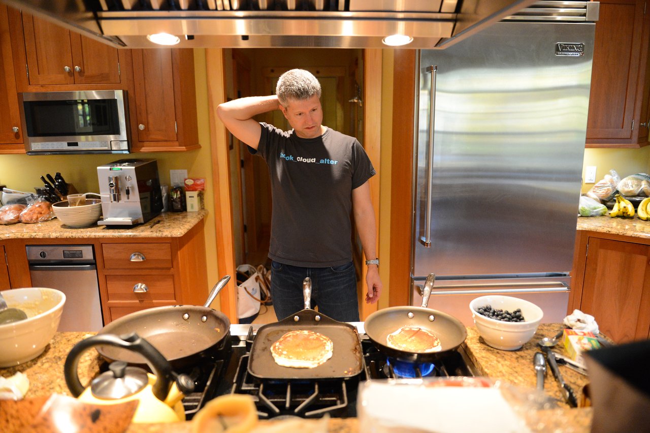 A man in a kitchen looks at pancakes cooking on a stovetop, scratching his head.