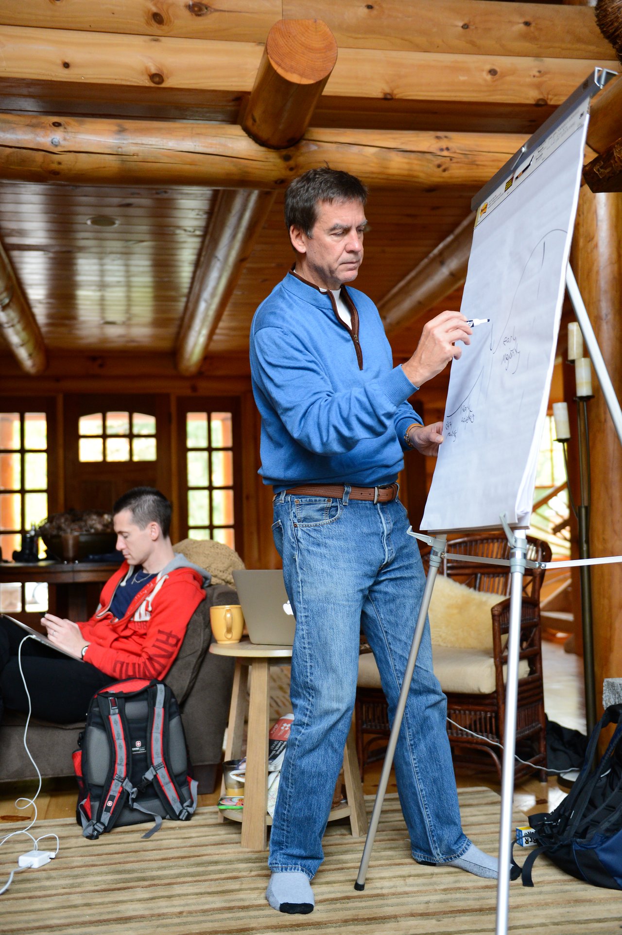 A man in a blue sweater writes on a flip chart while another person works on a laptop nearby.