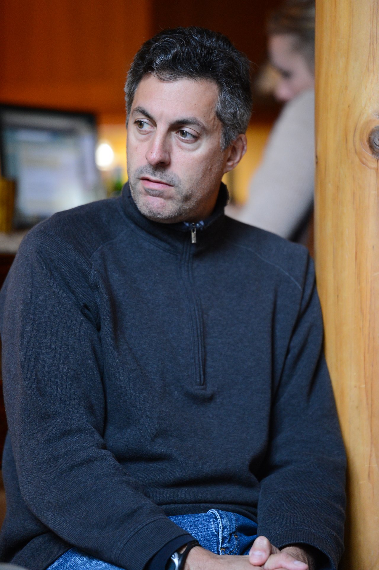 A man in a dark sweater sits indoors, looking to the side with a neutral expression.