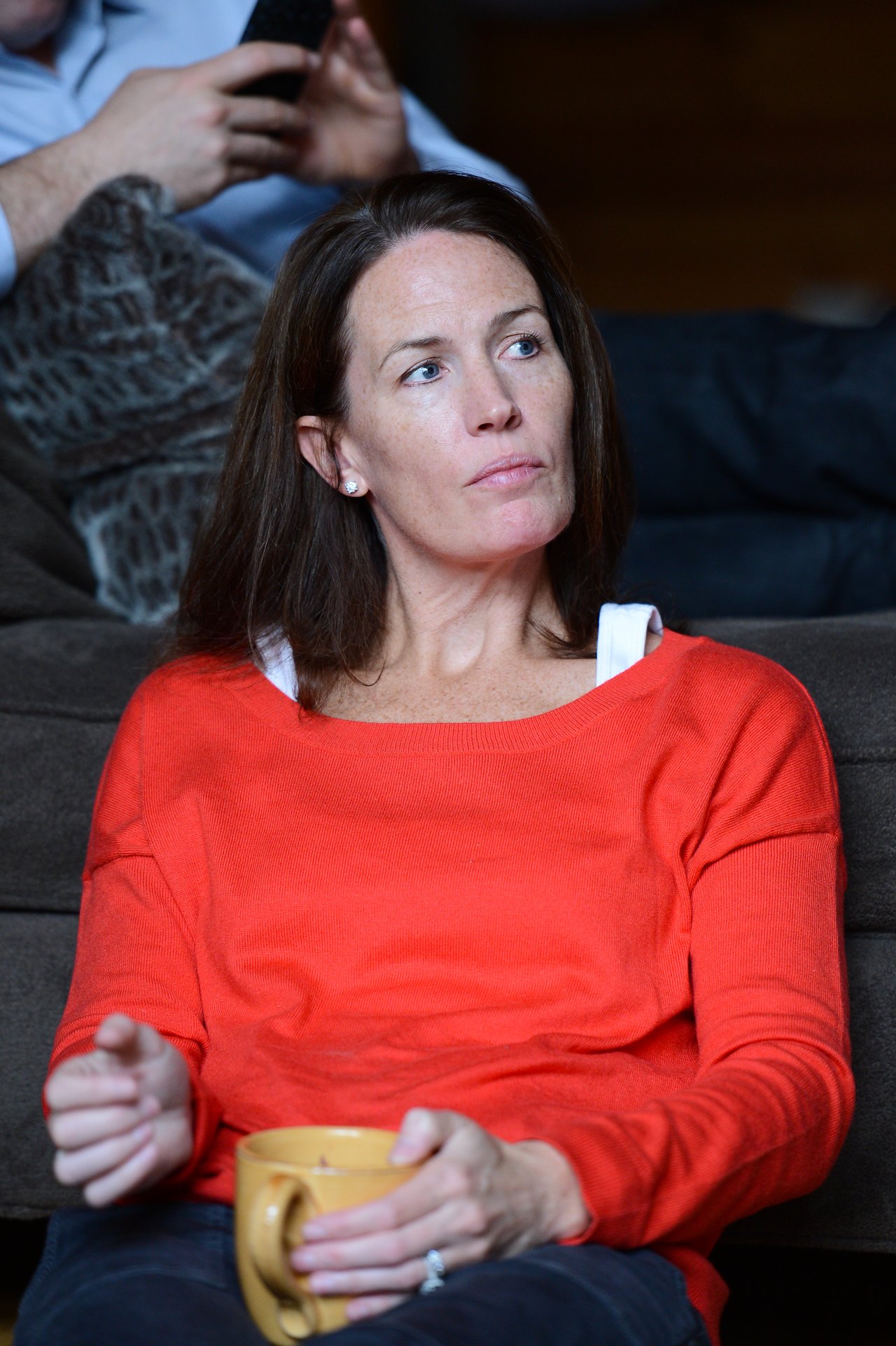 A woman in a red sweater sits on a couch, holding a yellow mug and looking to the side.