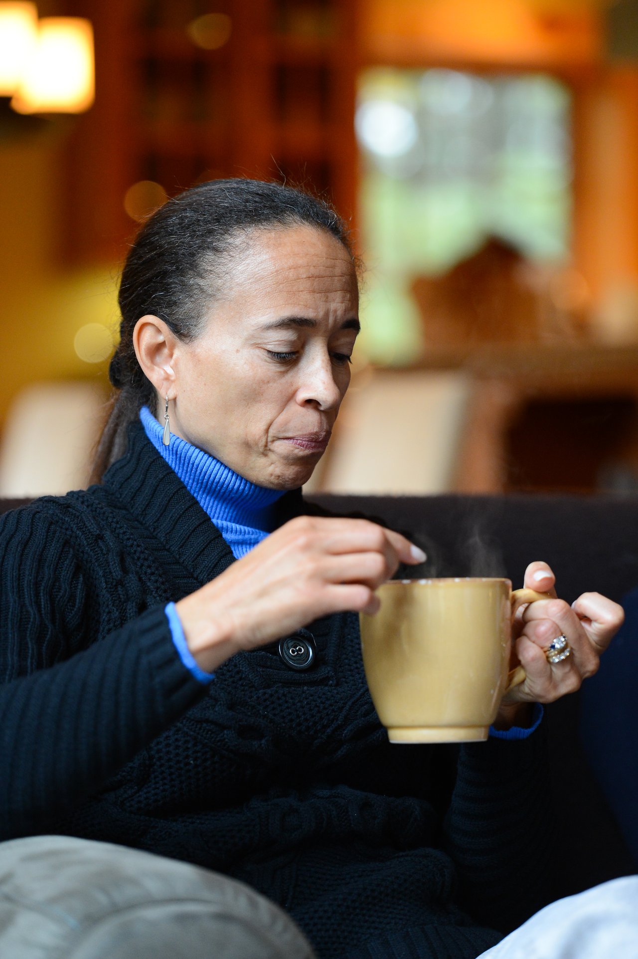A woman in a black sweater stirs a hot drink in a yellow mug while sitting indoors.