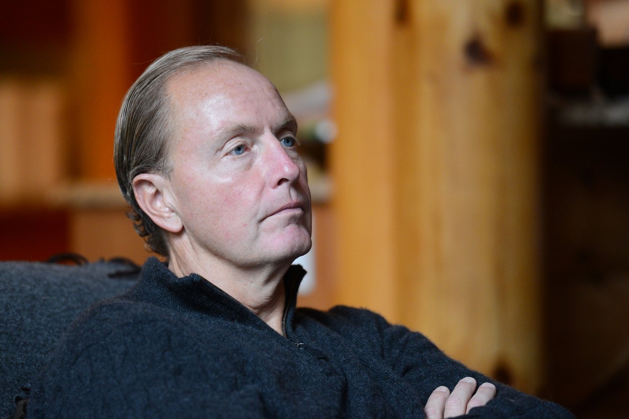 A man in a black sweater sits with arms crossed, attentively listening in a warmly lit indoor setting.