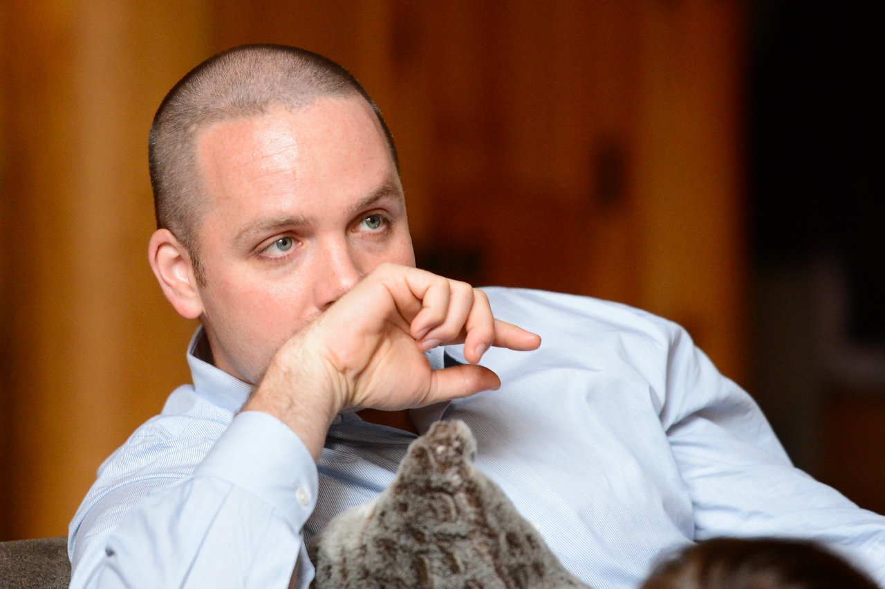 A man in a light blue shirt sits on a couch, resting his hand near his face, appearing thoughtful.
