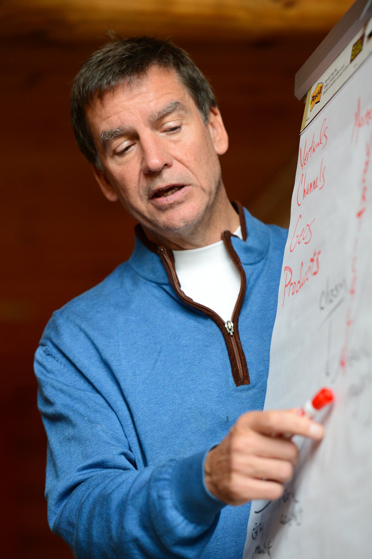 A man in a blue sweater writes on a flip chart with a red marker during a presentation.