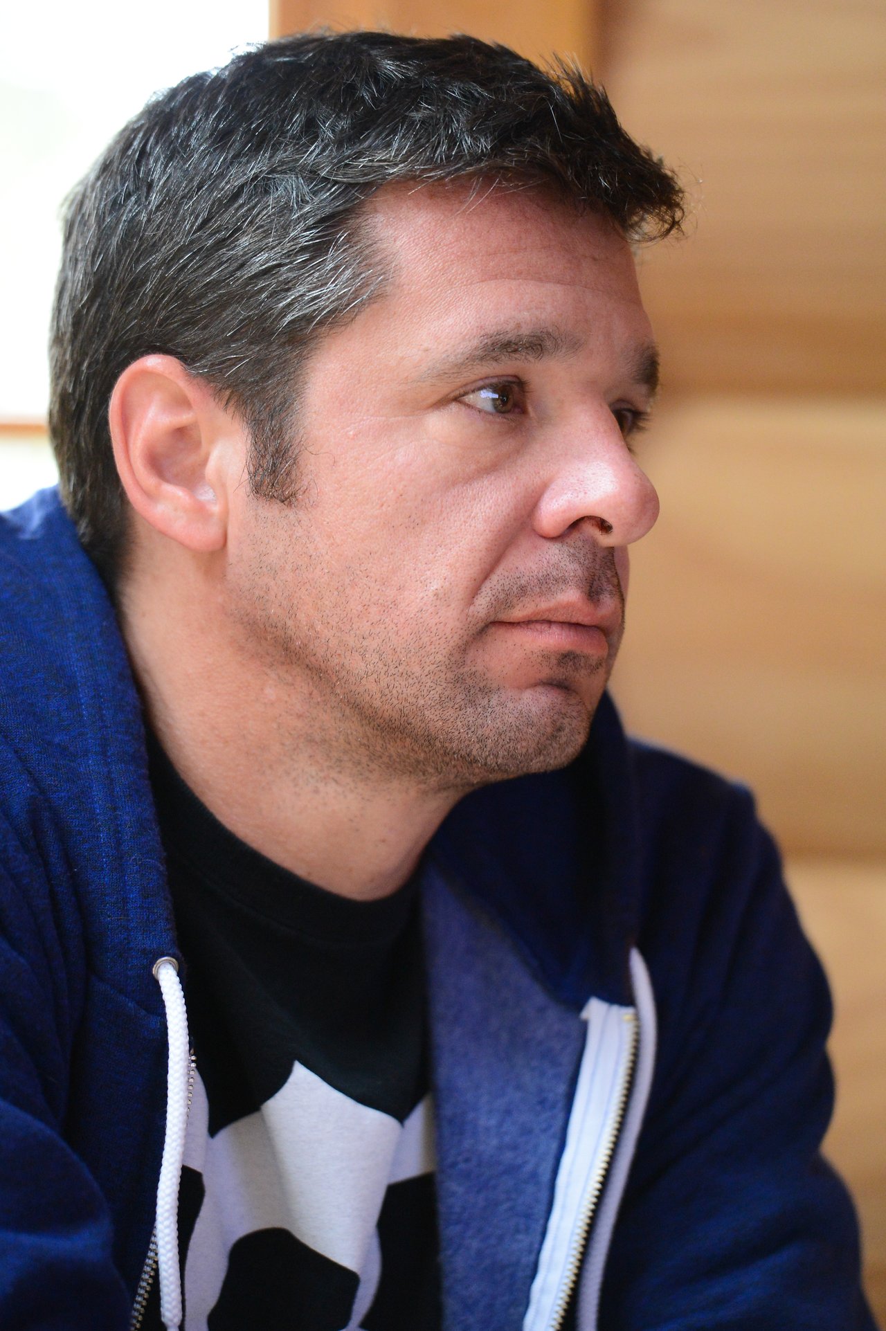 A man in a blue hoodie sits indoors, looking to the side with a focused expression.