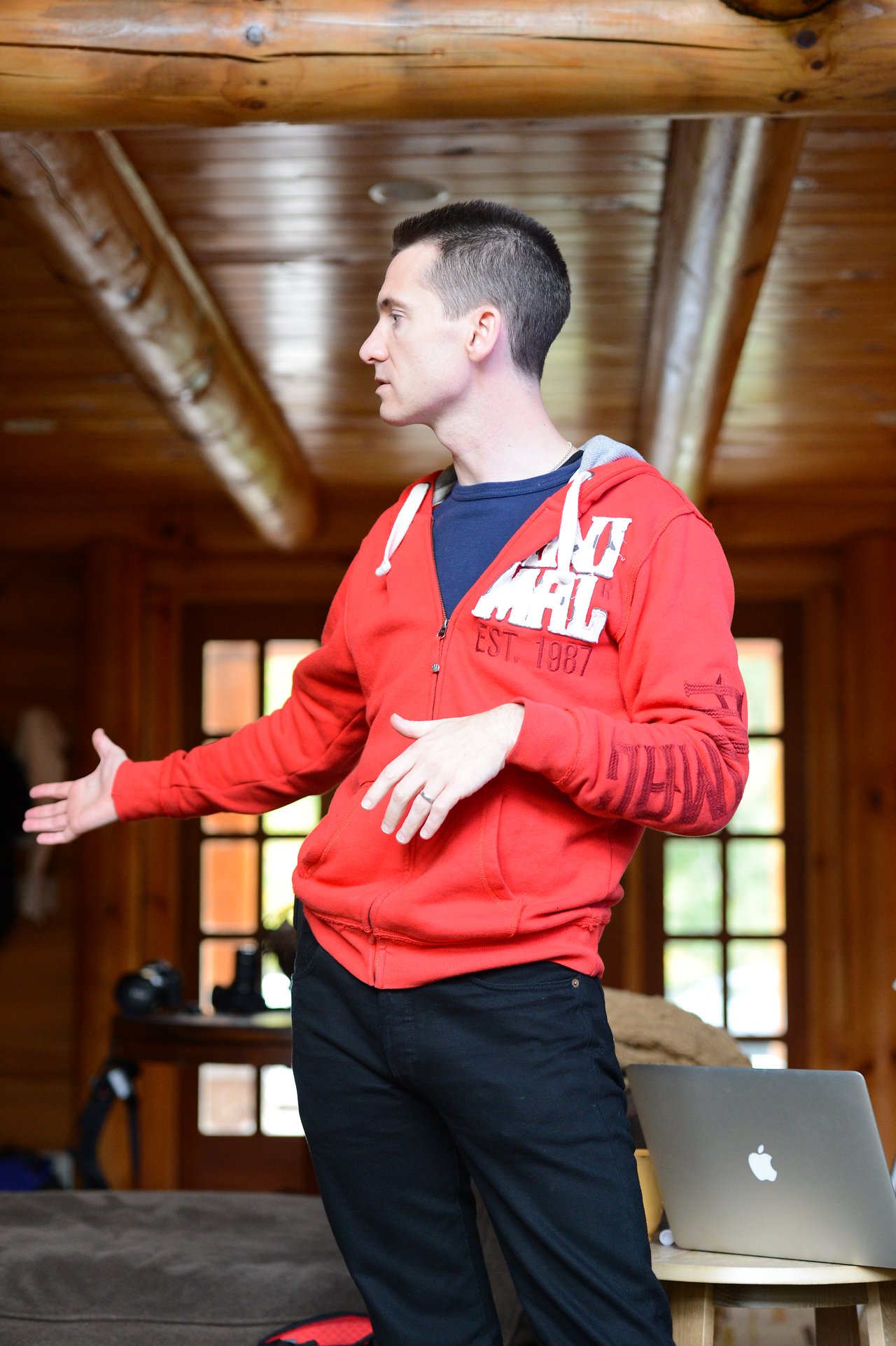 A person in a red hoodie gestures while speaking in a wooden cabin, with a laptop nearby.
