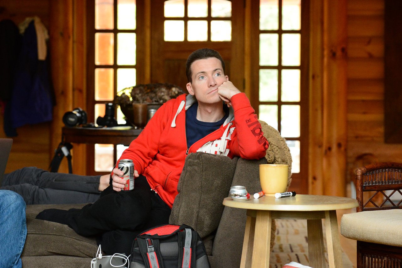 A person in a red hoodie sits on a couch, holding a soda can and resting their head on one hand.