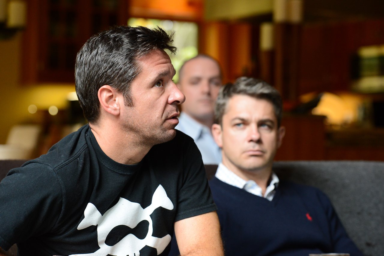 Three men sit indoors, attentively listening to something off-camera.