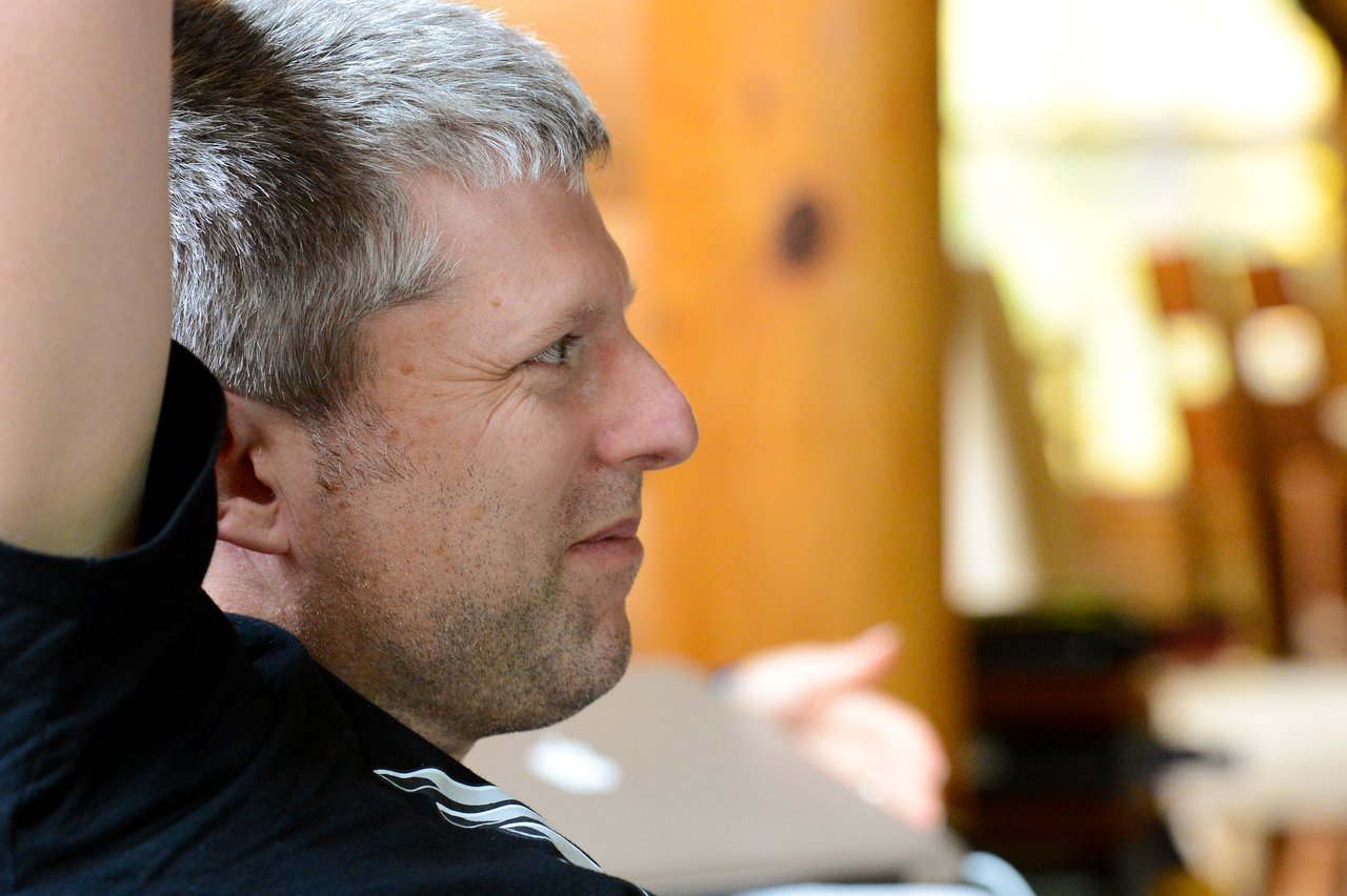 A man with short gray hair raises his arm while looking to the side in a casual setting.