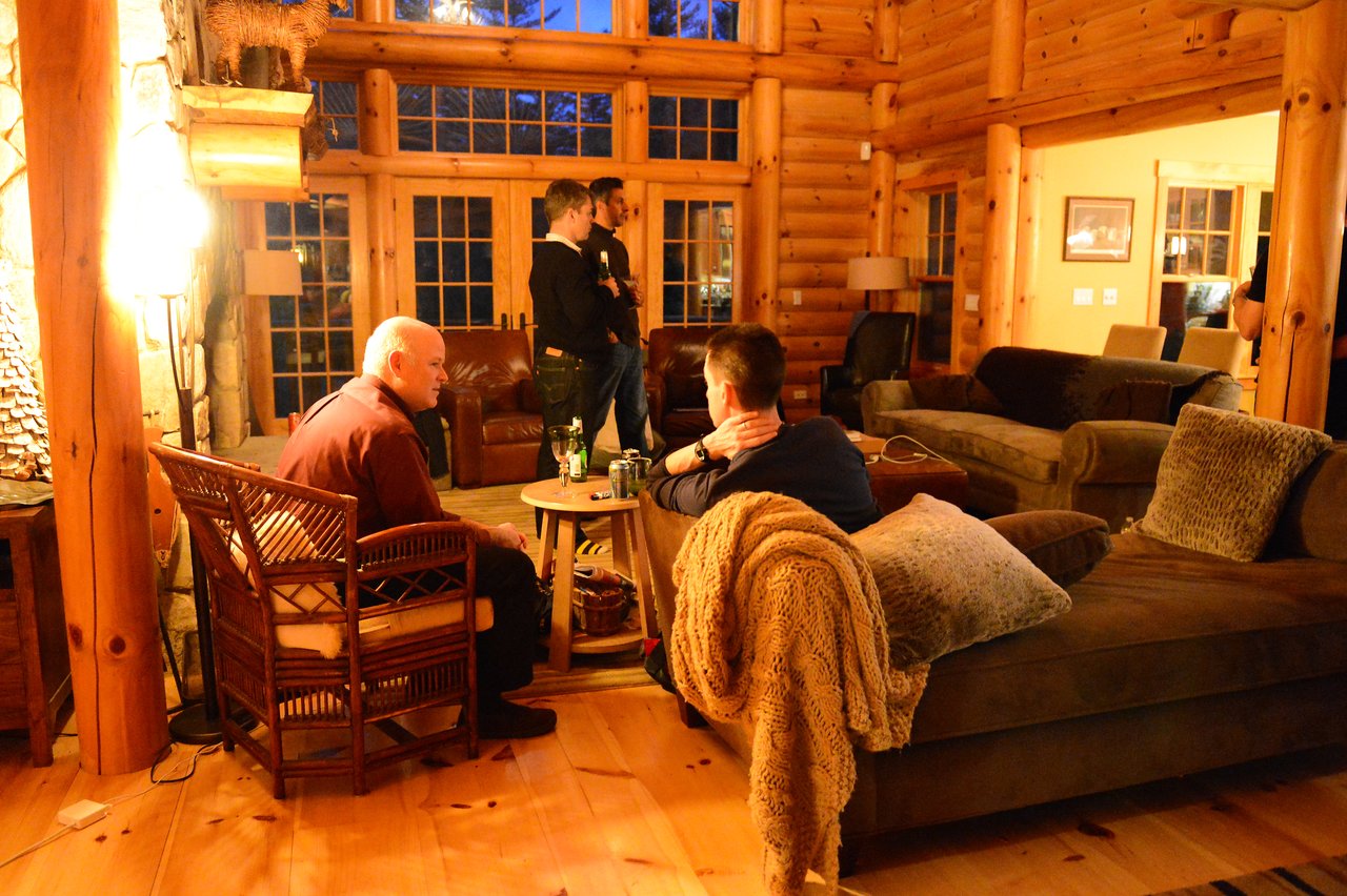 A group of people in a log cabin, engaged in conversation and relaxing with drinks.