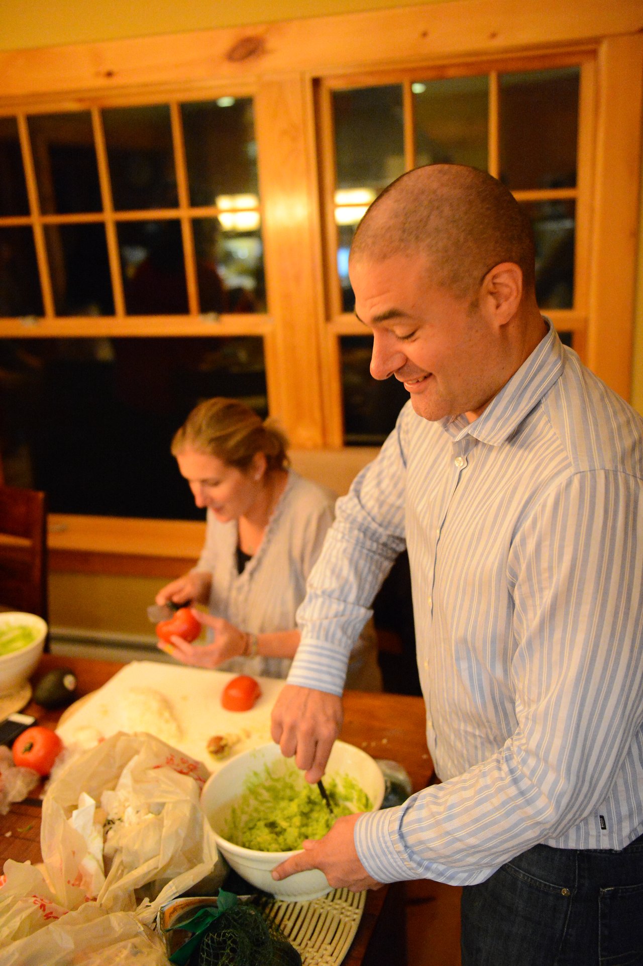 Two people prepare food in a kitchen; one mixes guacamole while the other chops tomatoes at a table.