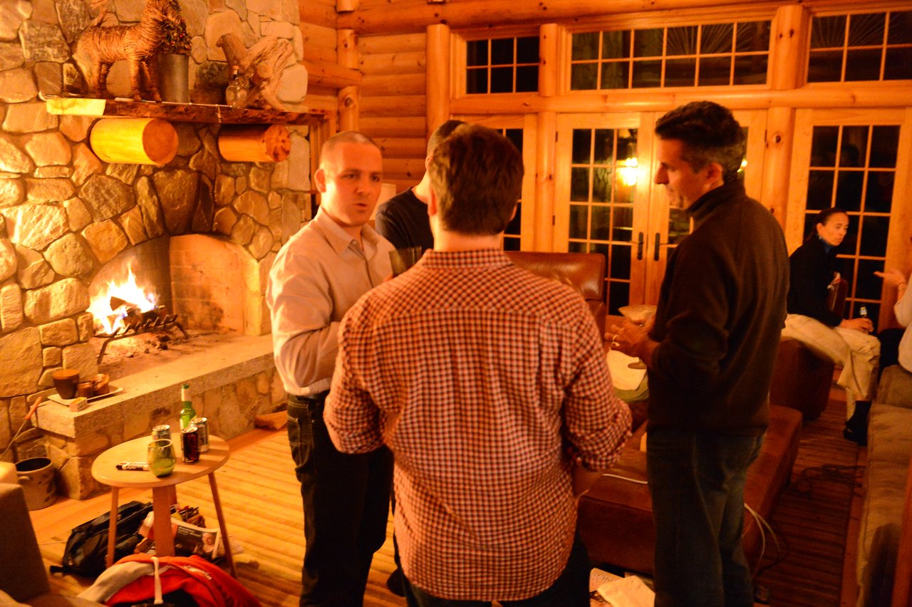 A group of people engaged in conversation in a cozy cabin setting with a fireplace in the background.
