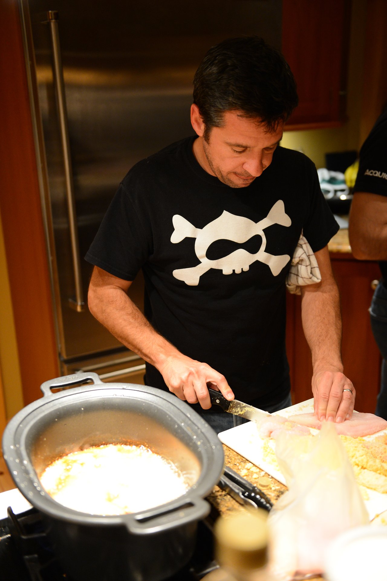 A man in a black t-shirt cuts raw chicken on a cutting board in a kitchen.