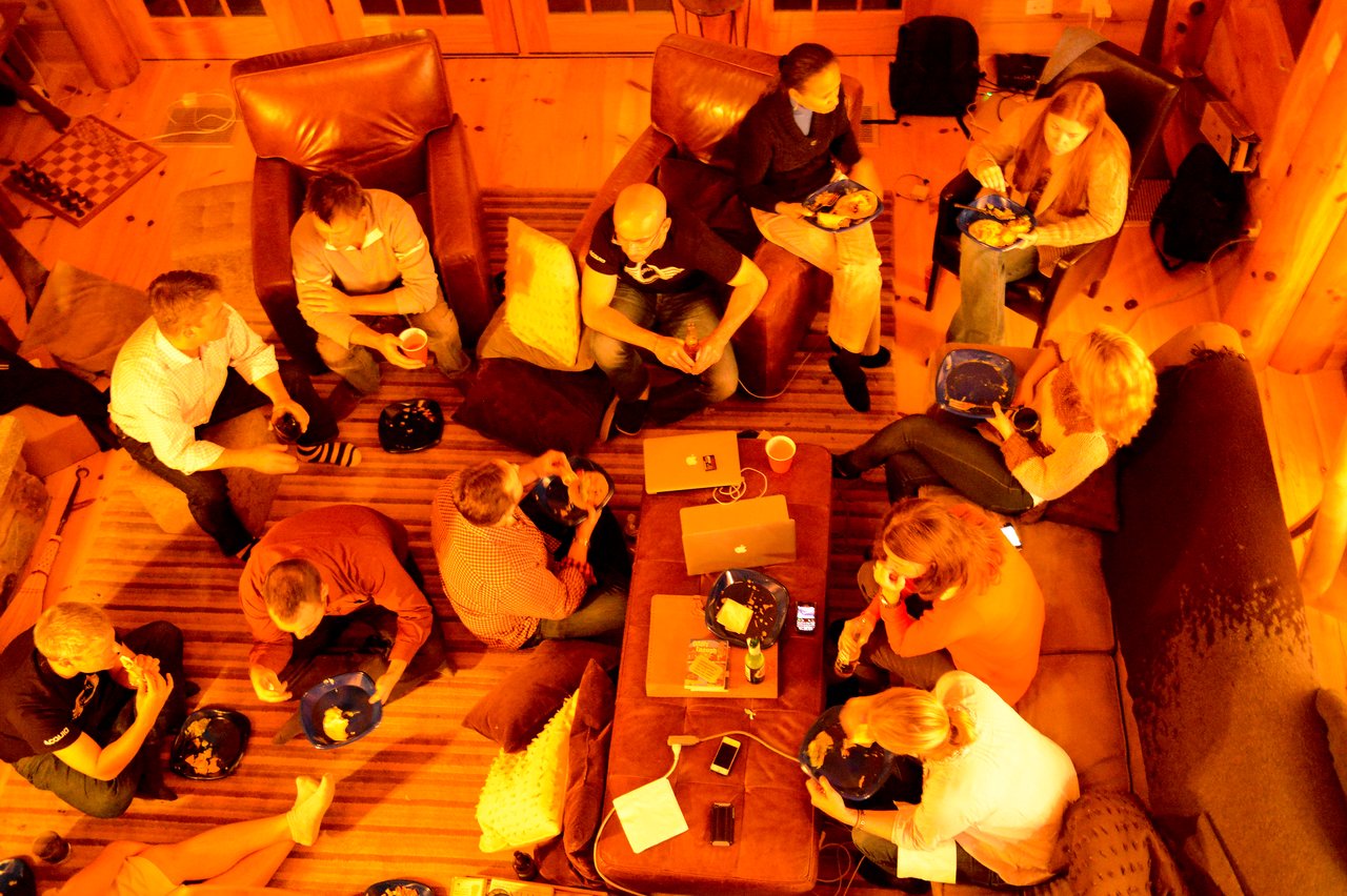 A group of people sits in a cozy room, eating and talking around a table with laptops and phones.