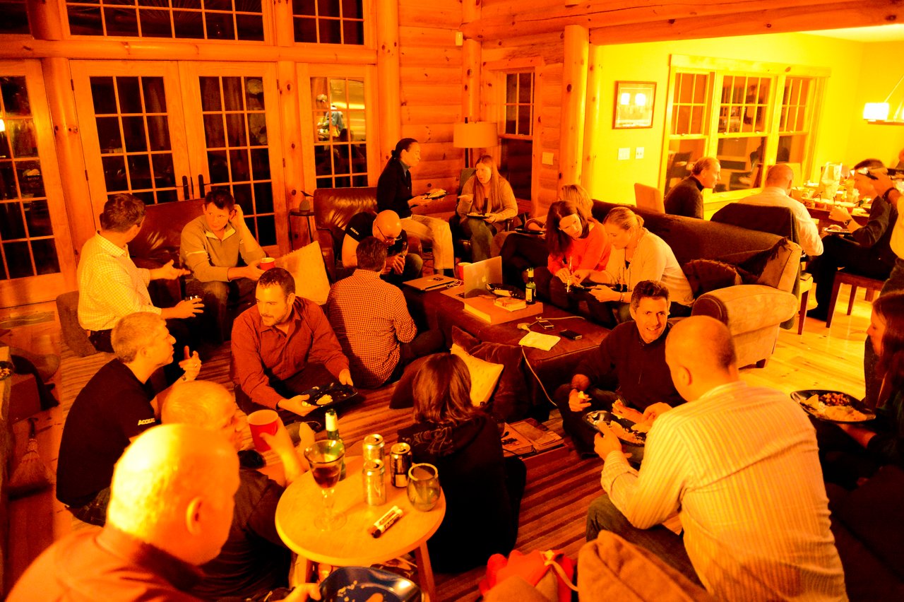 A group of people gathers in a cozy cabin, eating, drinking, and socializing during an offsite event.